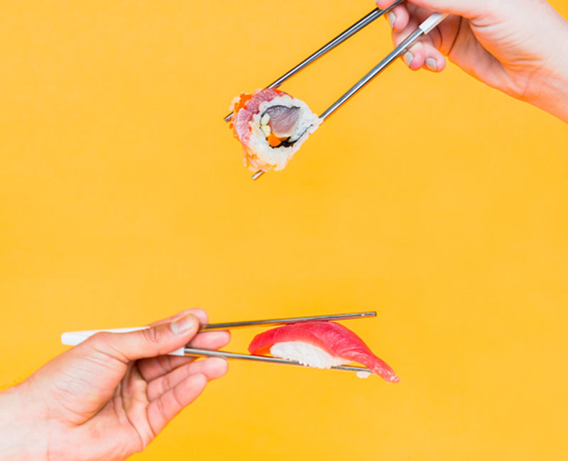 Two hands holding sushi with chopsticks against an orange background