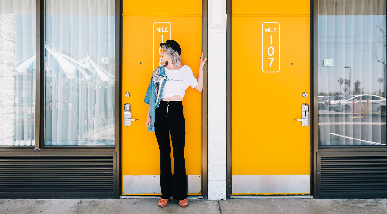 Girl in black pants standing in front of yellow motel door with mile marker sticker graphic