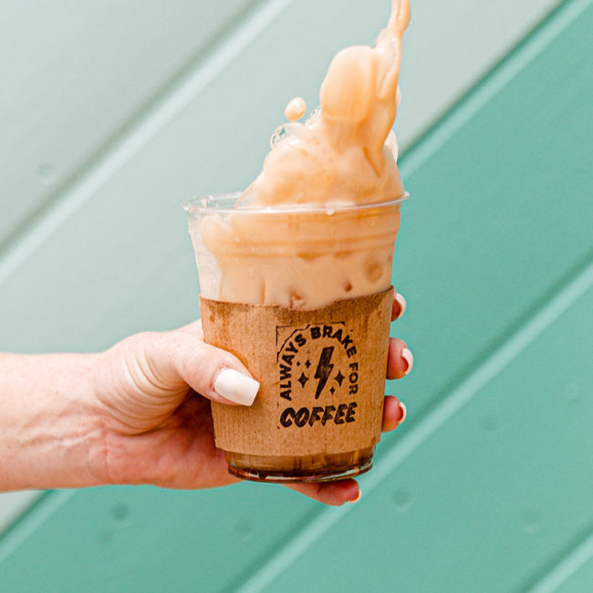 Hand holding an iced coffee that's spilling with a hand stamped sleeve that says "Always Brake for Coffee" against a light teal background